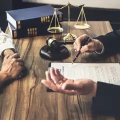 Prawnik omawia z klientem dokumenty, na stole młotek sędziowski, waga i księgi. Klient trzyma dłoń prawnika, podpisując list intencyjny.