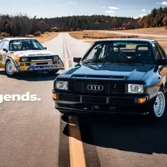 Dwa kultowe Audi S1 Quattro, jeden czarny, drugi w barwach rajdowych, stoją na pustej drodze.