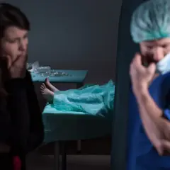 Smutna kobieta i lekarz w masce, obok zakryte ciało na stole. Kiedy sekcja jest obowiązkowa, emocje biorą górę.