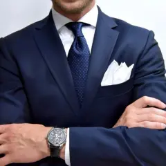 Elegancki mężczyzna w granatowym garniturze, białej koszuli i bordowym krawacie w kropki. Biała poszetka i zegarek dopełniają stylizacji.