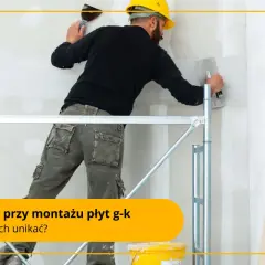 Fachowiec w żółtym kasku i roboczych spodniach na rusztowaniu wygładza ścianę z regipsu.