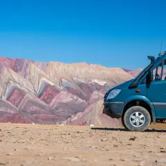 Un camping car tout terrain bleu est garé sur un sol rocailleux, avec en arrière-plan des montagnes aux couleurs vives et un ciel bleu éclatant.
