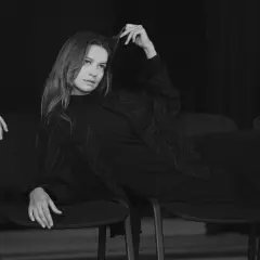 Kobieta siedzi na krześle, bawiąc się włosami. Jej spojrzenie jest nieobecne, jakby była duchem, upiorem z innego świata.