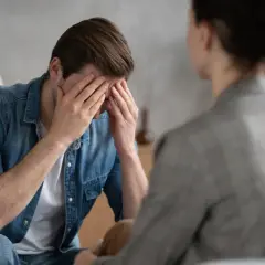 Mężczyzna w dżinsowej koszuli z rękami na twarzy, siedzący naprzeciwko drugiej osoby.