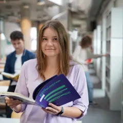 Studentka z uśmiechem trzyma broszurę, być może dotyczącą stypendium socjalnego. W tle inni studenci w bibliotece.