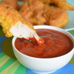 Chrupiący nugget zanurza się w gęstym, czerwonym sosie do nuggetsów. W tle widać talerz z innymi nuggetami.