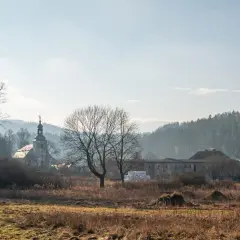 Krajobraz z kościołem i zabudowaniami, w tle las. W oddali widać mgliste wzgórza, charakterystyczne dla rudaw janowickich.