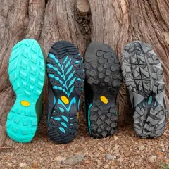 Four hiking boot soles lean against a tree. From left to right: bright teal, black with teal accents, black with circular lugs, and grey with a rugged tread. These sneaker soles are hard and slippery, designed for grip.