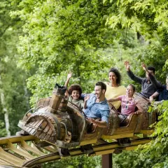Famille souriante et joyeuse sur un wagon de parc attraction, bras levés, profitant d'une descente dans un décor verdoyant.