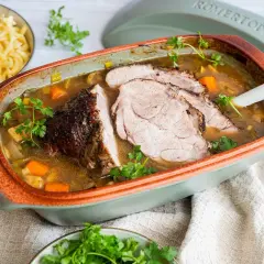 Saftiger schweinebraten im schmortopf, mit Karotten und Lauch, garniert mit Petersilie. Daneben eine Schüssel mit Spätzle.