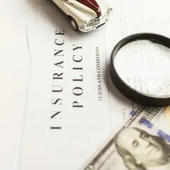 Model zabawki, lupa i dolarowe banknoty na dokumencie "Insurance Policy" sugerują analizę ubezpieczenia assistance.