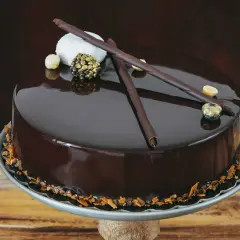 Torta cioccolatosa goduriosa con glassa lucida, decorata con riccioli di cioccolato, nocciole e una sfera dorata.