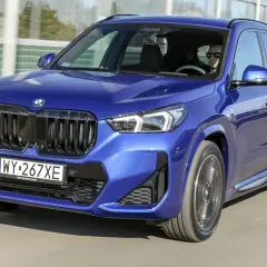 Niebieskie BMW X1 w ruchu drogowym. Kierowca w okularach przeciwsłonecznych. Zbliżenie na przód auta, widoczne logo BMW i atrapa chłodnicy. Wymiary BMW X1 pozwalają na komfortową jazdę.
