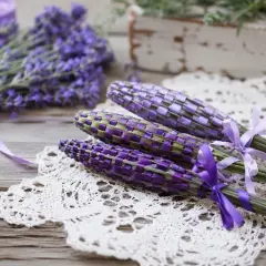 Drei geflochtene Lavendel-Stäbe mit lila Bändern auf einer Spitzendecke. Im Hintergrund ein Bund frischer Lavendel.