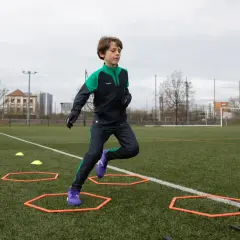 Chłopiec w zimowym stroju piłkarskim ćwiczy na boisku, pokonując tor przeszkód. Idealny przykład, jak ubrać dziecko na trening piłkarski zimą.