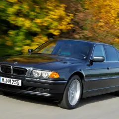 Ciemnoszary BMW E38 Long sunie po drodze, otoczony jesiennymi drzewami w pełnym kolorze.