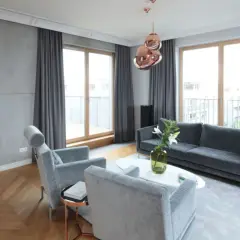 Nowoczesny salon z betonową ścianą, drewnianą podłogą w jodełkę, szarymi meblami i miedzianymi lampami. Lamele i beton tworzą industrialny klimat.