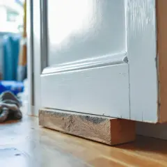 Un bloc de bois soutient une porte blanche pour la raboter. Des gants de travail sont posés au sol.