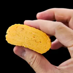 A hand holds a slice of golden-yellow dashimaki tamago, revealing its layered, fluffy texture.