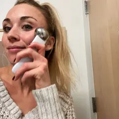 Kvinna med blont hår använder en vit och silverfärgad **ice roller ansikte** för att massera kinden.