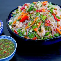 En stor skål med färgglad glasnudel sallad med räkor, paprika och grönsaker. Serveras med en dressing och hackade nötter.