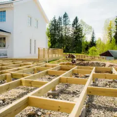 Grundläggande stomme för att bygga veranda framför ett vitt hus. Träbjälkar ligger på grus och stenar, redo för nästa steg.