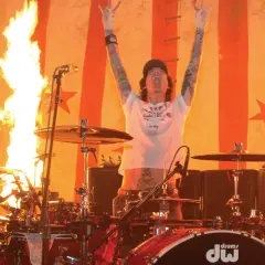 Tommy Lee w ogniu, z rękami w górze i pałeczkami perkusyjnymi, na tle płonącej sceny.