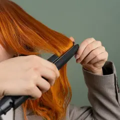 Kobieta prostuje rude włosy prostownicą.