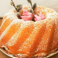 Puszysta babka jogurtowa, oprószona cukrem pudrem, ozdobiona bazieczkami i różowymi kwiatami. Idealna na Wielkanoc.