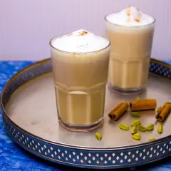 Två glas med krämig chai latte, toppad med skum och kanel. Kardemumma och kanelstänger på brickan ger en hint om detta chai latte recept.