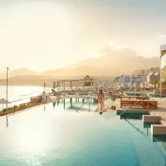 En kvinna i en blommig klänning går mot en infinitypool vid havet på Kreta, Makrigialos. Solnedgången färgar himlen gyllene.