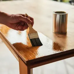 Main d'un artisan appliquant un vernis pour restaurer une table en contreplaqué. La finition brillante redonne vie au bois.