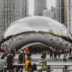 A crowd gathers around Cloud Gate, a reflective public art installation in Chicago, mirroring the city's skyscrapers and sky.
