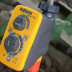 Ein gelber Hozelock Bewässerungscomputer mit Drehreglern für Frequenz und Minuten, angeschlossen an einen Wasserhahn.
