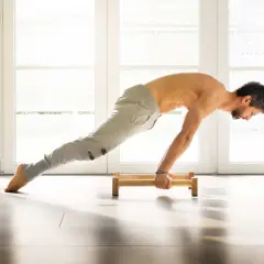 Hombre haciendo flexiones en paralelas de madera en un suelo claro, con luz entrando por ventanas al fondo.