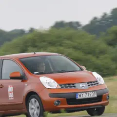 Pomarańczowy Nissan Note E12 z kobietą za kierownicą jedzie po drodze wśród zieleni.