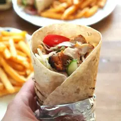 Ręka trzymająca prawdziwy kebab z kurczakiem, warzywami i frytkami w tle.