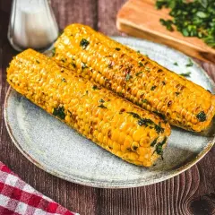 Zwei goldgelbe Maiskolben, perfekt gegrillt und mit Kräutern bestreut, liegen auf einem Teller. Ein Genuss für alle, die Maiskolben grillen lieben!