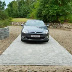 Grå bil parkerad på en vacker betong uppfart. Omgiven av grönskande träd och en stenmur.