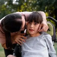Ciepły uścisk mamy dla dziecka z zespołem Retta. Wózek inwalidzki i zieleń w tle.