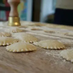 Świeżo przygotowane domowe ravioli na drewnianym blacie, posypane mąką. W tle widoczny przyrząd do wycinania pierogów.
