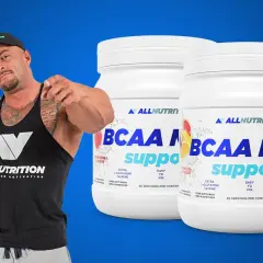 Muskularny mężczyzna w czapce i koszulce Allnutrition wskazuje palcem na dwa słoiki suplementu BCAA MAX.