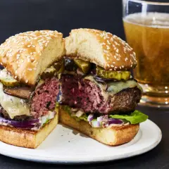 Przekrojony soczysty burger z serem, ogórkiem i grzybami, obok szklanka piwa.