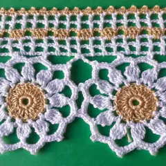 Delicati merletti uncinetto bianchi e dorati, con motivi floreali e geometrici, creano un bordo decorativo su sfondo verde.