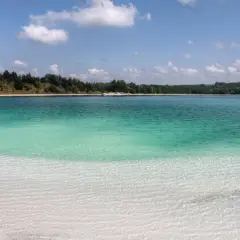 Lazurowe jezioro z piaszczystą plażą i lasem w tle. Woda jest krystalicznie czysta, a dno widoczne.