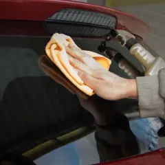 Ręka z pomarańczową mikrofibrą wyciera tylną szybę czerwonego samochodu. Obok widać dyszę spryskiwacza, sugerującą myjnię automatyczną bezdotykową.