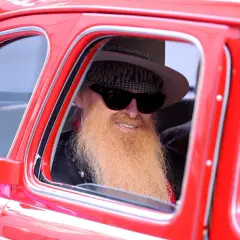 Billy Gibbons, jeden z członków ZZ Top, w stylowym kapeluszu i z charakterystyczną brodą, siedzi w czerwonym samochodzie.