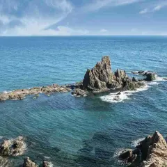 Formaciones rocosas emergen del mar turquesa en la **sierra de Cabo de Gata**. El sol ilumina las aguas cristalinas y el cielo azul con nubes blancas.