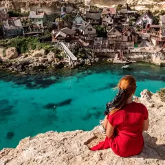 Widok na maltańską wyspę Popeye Village, gdzie turystka w czerwonej sukience podziwia turkusowe morze.