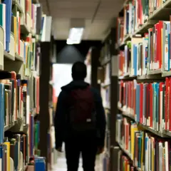 Ein Schüler mit Rucksack geht durch eine Bibliothek. Vielleicht sucht er nach Büchern, die ihm zeigen, dass das Gymnasium viel schwerer als Realschule ist.